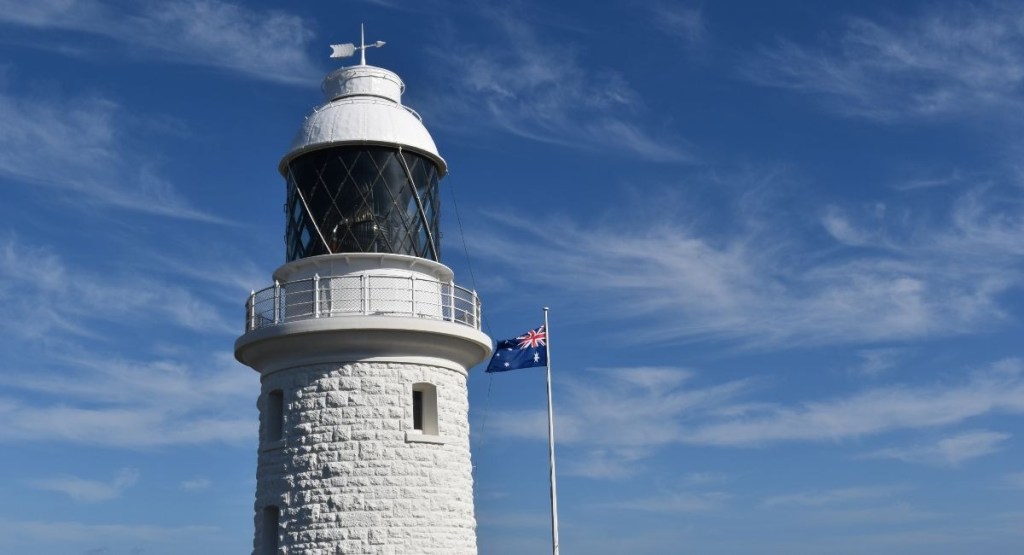 A tall, white lighthouse stands against a cloudy blue sky next to a waving flag. The lighthouse is constructed from white stone with a distinctive, paneled, glass light at the top, and the Australian flag waves from a tall pole to the right of the lighthouse.