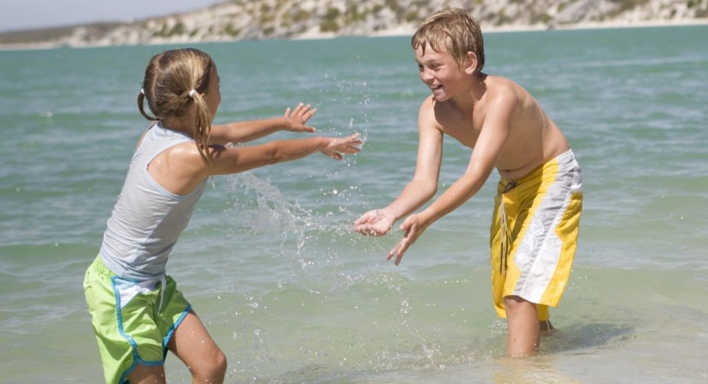 A girl and a boy with blonde hair are playing in the ocean on a sunny day. The children are waist-deep in the clear turquoise water and facing one another.