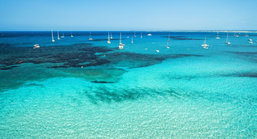 A high-angle shot reveals a stunning coastal view of turquoise waters on a clear, sunny day near Busselton, WA. Several boats are moored on the serene ocean surface.